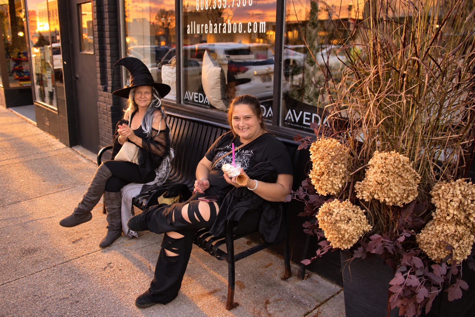 Two visitors in witch costumes enjoying a break on a bench beside autumn flowers