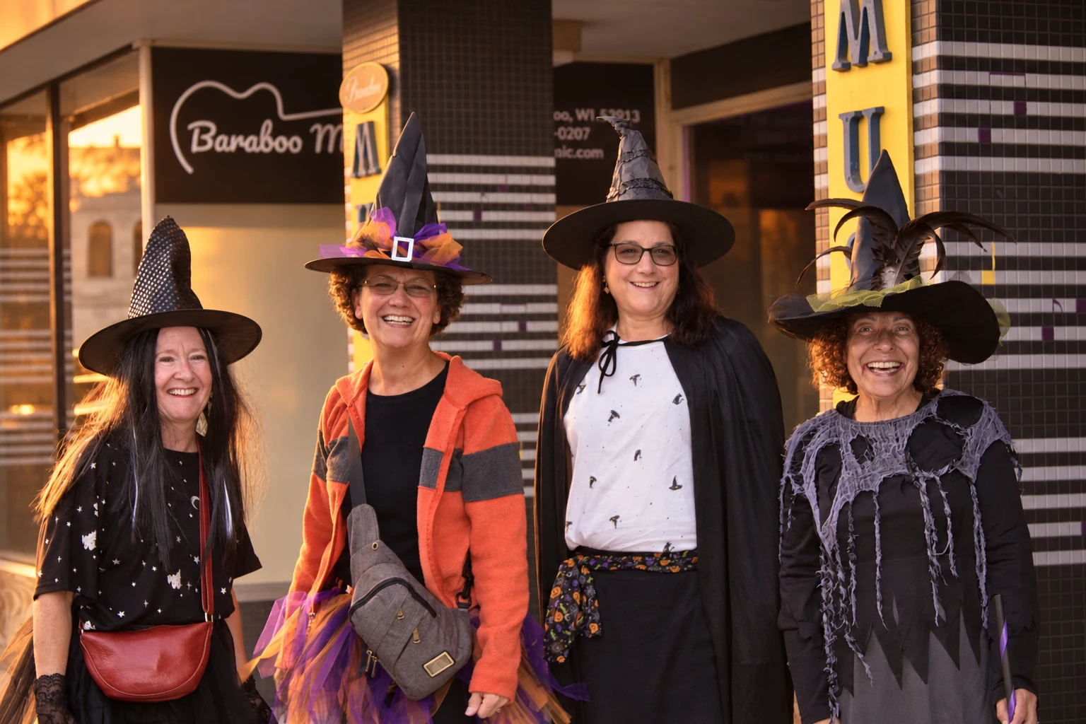 Four smiling visitors in decorated witch hats outside Baraboo Music