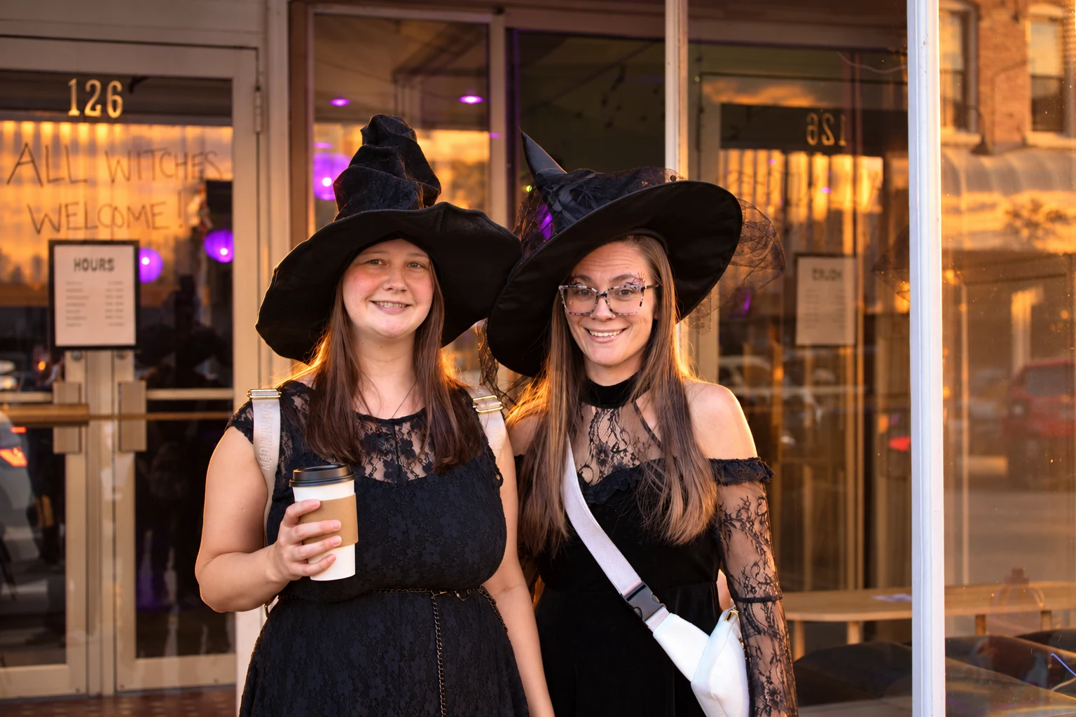 Two friends in black costumes and witch hats posing outside a shop