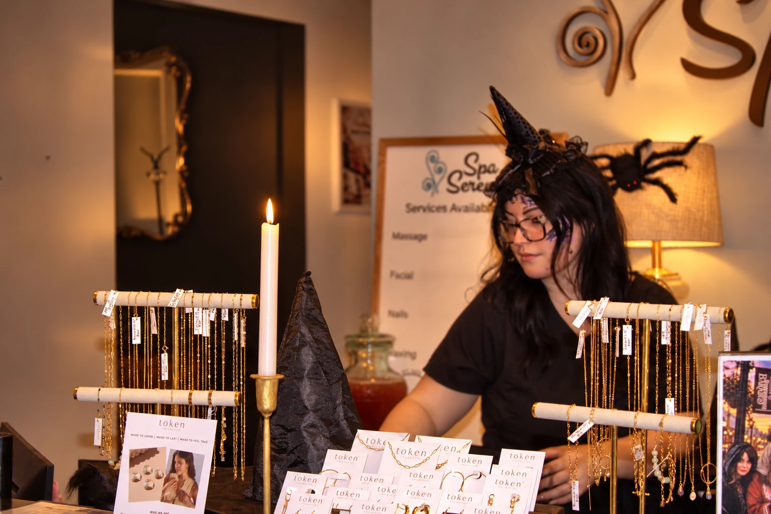 A vendor in a small witch hat behind jewelry displays at Spa Serenity