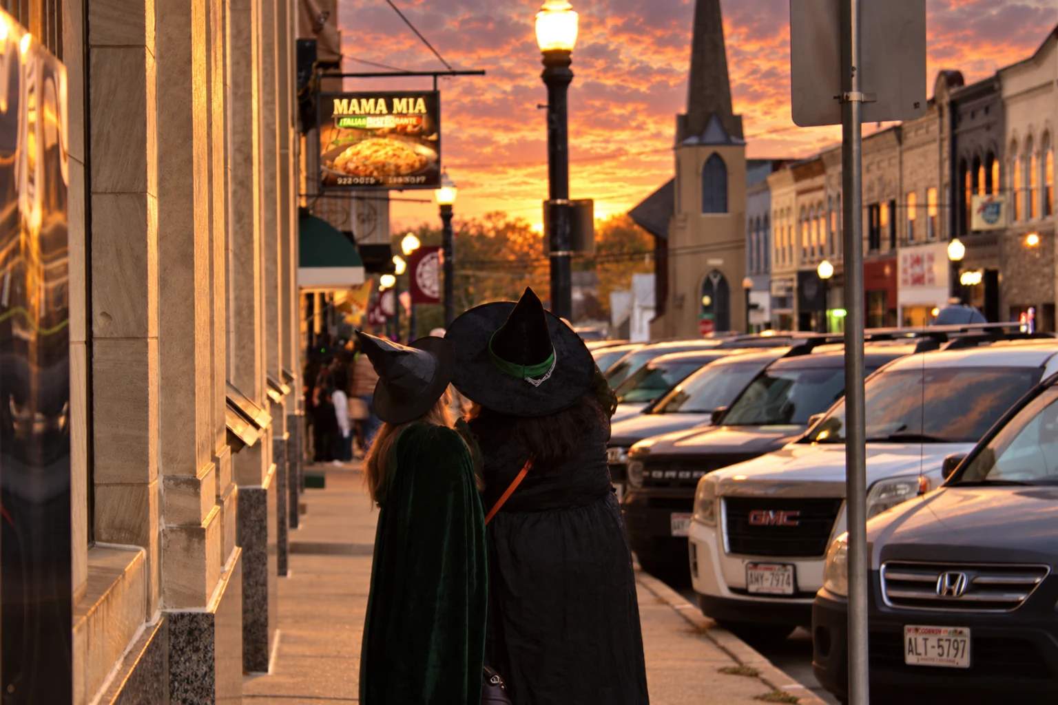 Two visitors in witch hats walking away past storefronts and parked cars